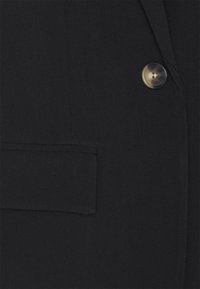 Détail d’un manteau noir montrant un seul bouton brunâtre et une poche à rabat horizontale sur le côté gauche.