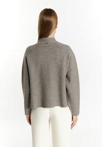 Pulover gri cu guler înalt, mâneci raglan și croială relaxată. Textură netedă cu tiv despicat, purtat peste pantaloni albi.