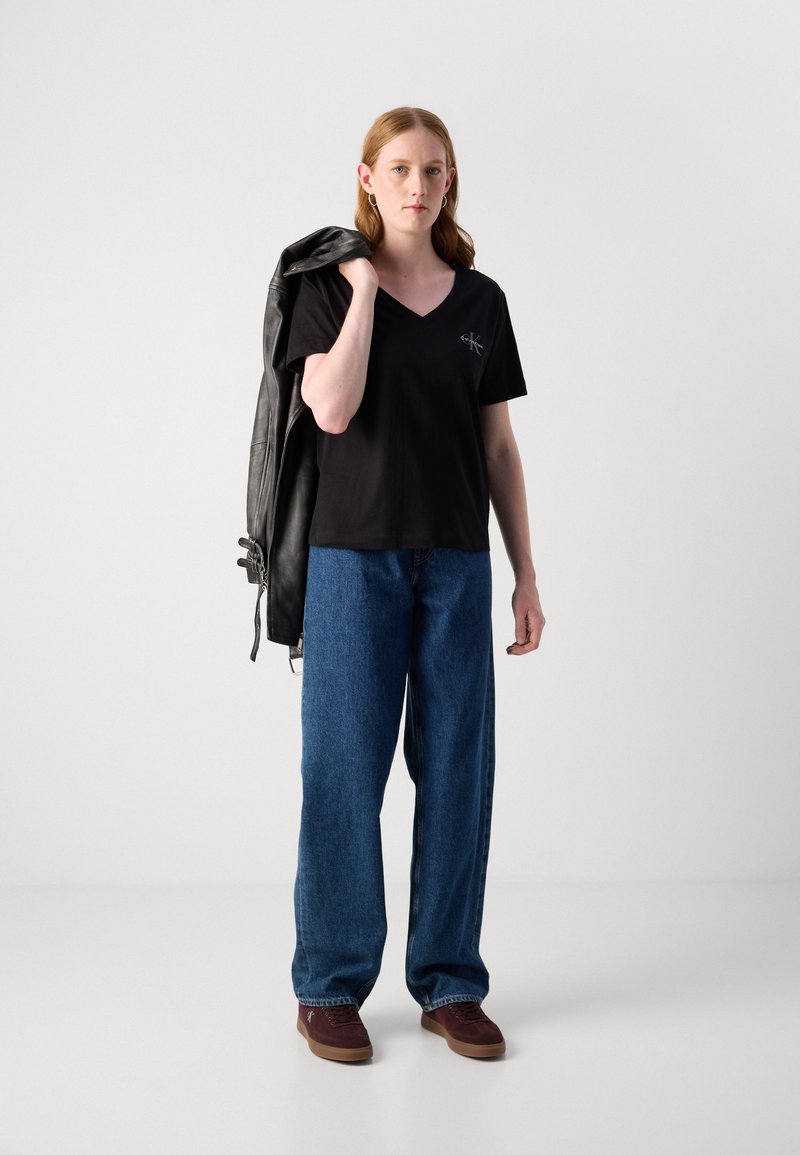 Czarna koszulka z dekoltem w serek z logo na piersi, połączona z szerokimi, niebieskimi dżinsami i brązowymi trampkami. Skórzana kurtka swobodnie narzucona na ramię.