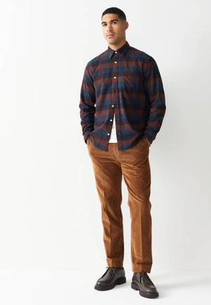 Košeľa na gombíky v tmavomodrom a hnedom károvanom látke, kombinovaná s hnedými manšestrákovými nohavicami a čiernymi koženými topánkami. Minimalistický štýl, bez vzorov.