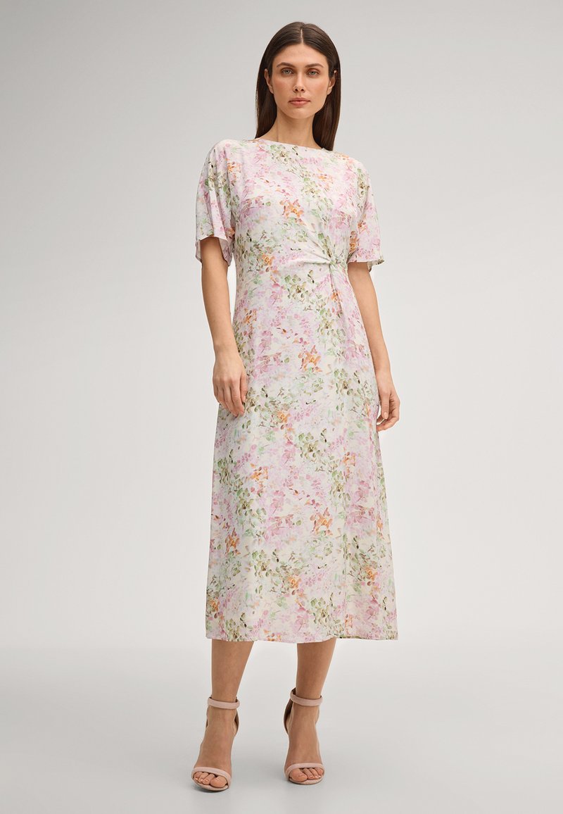 Donna in piedi con un vestito midi a fiori pastello con maniche corte, dettaglio a nodo in vita e tacchi nudi con cinturino alla caviglia su sfondo neutro.