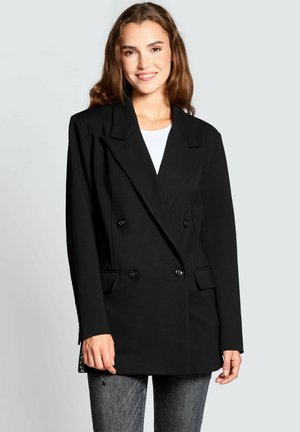 Schwarze zweireihige Blazerjacke mit breiten Revers, ausgestattet mit zwei Vordertaschen und schwarzen Knöpfen; kombiniert mit einem weißen Oberteil und grauen Jeans.