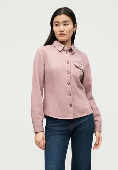 Νεαρή γυναίκα με μακριά σκούρα μαλλιά, που φοράει ανοιχτό ροζ πουκάμισο με τσέπη στο στήθος και σκουρόχρωμο μπλε τζιν, μπροστά σε απλό φόντο.