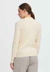 Maglione a maglia cable color crema con maniche lunghe e orlo a coste. Texture liscia e design aderente con dettagli sottili lungo la superficie.