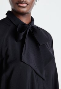 Blouse noire avec un col en nœud papillon proéminent, en tissu lisse, avec un léger éclat et une coupe décontractée. Vue rapprochée mettant en avant les détails du design.