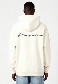 Felpa color crema con cappuccio regolabile. Presenta la scritta nera ricamata "Amour" sul retro. Polsini e orlo a coste. Design casual.
