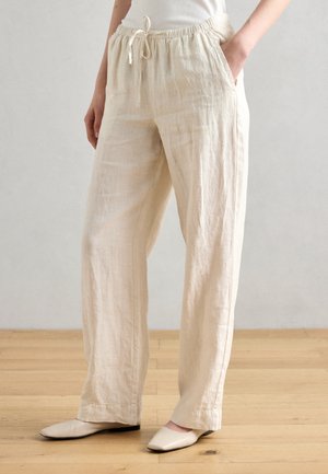 Personne portant un pantalon ample en lin beige avec une taille élastique et un cordon, les mains dans les poches, debout sur un sol en bois avec des chaussures beiges.