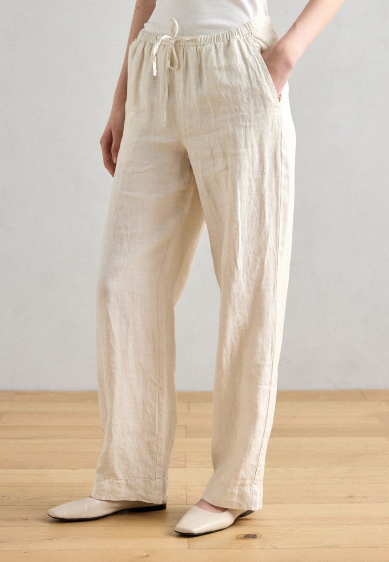 Personne portant un pantalon ample en lin beige avec une taille élastique et un cordon, les mains dans les poches, debout sur un sol en bois avec des chaussures beiges.