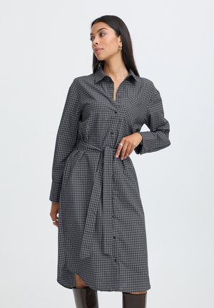 Femme portant une robe-chemise à manches longues grise à carreaux avec ceinture à la taille, debout, une main sur la hanche et regardant sur le côté.