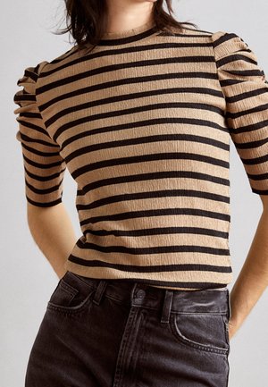 Femme portant un haut texturé beige et noir à rayures horizontales avec des manches courtes bouffantes et un jean noir taille haute.