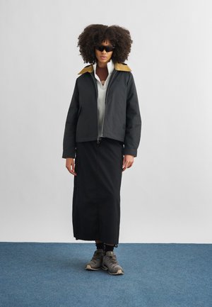 Kihara juustega naine, kandes musti päikeseprille, musta jopet tan-värvi kraega, valget lukuga pusa, pikka musta seelikut ja halli vabaajajalatseid, seisab sinisel põrandal.