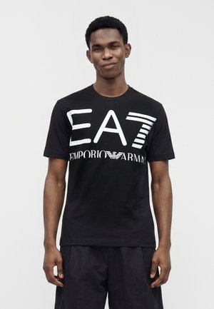 Άνδρας που φοράει μαύρο t-shirt Emporio Armani EA7 με μεγάλο λευκό λογότυπο, στέκεται μπροστά σε απλό ανοιχτό γκρι φόντο.