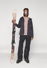 Hiihtoasu koostuu mustasta takista, jossa oransseja yksityiskohtia, vaaleanpunaisesta kuvioidusta neuleesta, mustista housuista sekä suksista, joissa on siteet. Suksissa on graafinen kuvio.