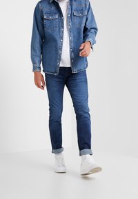 Chaqueta de denim en azul claro con dos bolsillos en el pecho, llevada sobre una camiseta blanca. Jeans de color azul oscuro y zapatillas blancas a la vista, con los puños remangados.