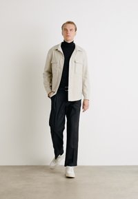Svijetlosmeđi košulja-jakna, crni turtleneck, široke crne hlače i bijele tenisice. Dva džepa na prsima jakne i minimalistički stil.