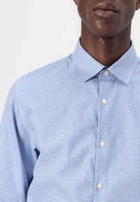 Sinivalkoruudullinen nappipaita, jossa on jämäkkä kaulus ja hienovarainen tekstuuri, esitetty rintakehän yläpuolelta alkaen.