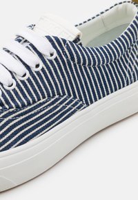 Modré a bílé pruhované plátěné tenisky s bílými tkaničkami, gumovou podrážkou a hladkým povrchem. Obsahují logo na jazyku a očkách.