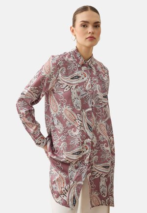 Vrouw draagt een overhemdjurk met lange mouwen, knopen en een roze, witte en zwarte paisley-print, handen in de zakken.