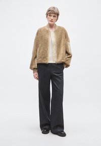 Jachetă bomber bej în stil Teddy, cu material texturat, manșete rib și buzunare laterale cu fermoar în față, asortată cu pantaloni evazați din denim închis la culoare.