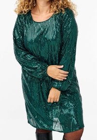 Grön paljettklänning med rundad halsringning, långa ärmar och lös passform, med en texturerad yta och glittrande detaljer genom hela.