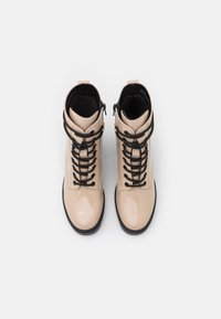 Bottes cheville en cuir verni beige avec lacets et accents noirs. Présentent une surface brillante, une fermeture éclair et un design à bout arrondi.