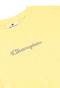 Camiseta de algodón amarilla de cuello redondo con un logotipo azul bordado en el pecho. Textura suave y corte estándar.
