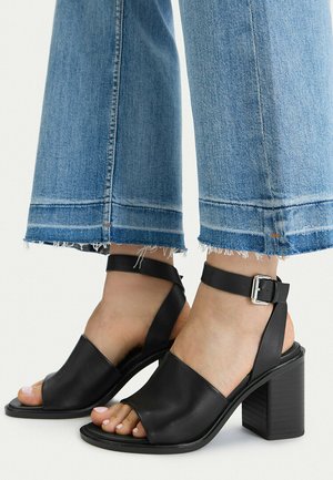 Piedi con sandali neri con tacco a blocco e punta aperta, con cinturini alla caviglia, abbinati a jeans azzurri corti con orli sfrangiati.