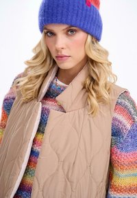 Beżowa pikowana kamizelka bez rękawów z jasną podszewką, noszona na kolorowym swetrze z dzianiny. Modelka ma na sobie fioletową dzianinową czapkę z czerwonym akcentem.