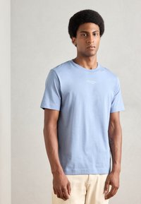 Tricou din bumbac de culoare albastru deschis, cu guler rotund, mâneci scurte și un mic logo alb impriminat pe piept. Textură netedă și croială lejeră.