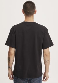 Camiseta de algodón negra con mangas cortas, ajuste relajado y cuello redondo. Tatuaje notable visible en el antebrazo. Vista posterior mostrada.