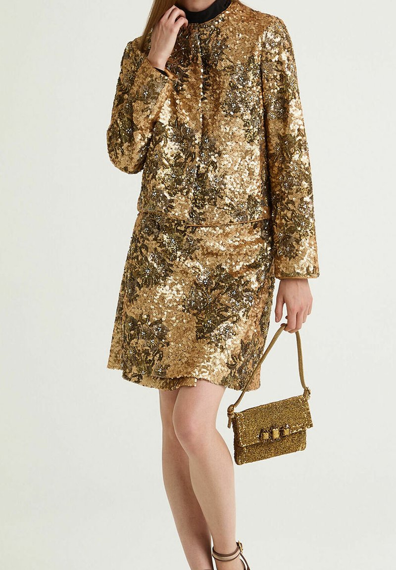 Ensemble twin en sequins dorés comprenant un haut à manches longues et une jupe. La tenue est accompagnée d'un petit sac à main assorti. Texture lisse et finition brillante.