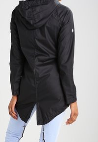 Veste noire à capuche avec un ourlet arrière plus long, matériau léger et détails cousus sur les épaules. Comprend un accent de logo sur la manche.