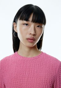 Rosa texturiertes Oberteil mit rundem Halsausschnitt und einem dezenten wellenförmigen Saum. Das Model trägt schlichte Creolen vor einem schlichten weißen Hintergrund.