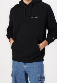 Personne portant un hoodie noir Lyle & Scott avec une poche avant et un pantalon cargo en denim bleu, les mains dans la poche du hoodie.