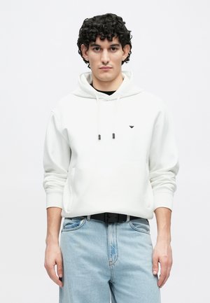 Witte hoodie met een voorvak en trekkoorden, voorzien van een klein zwart logo. Gecombineerd met lichtblauwe jeans en een zwarte riem.