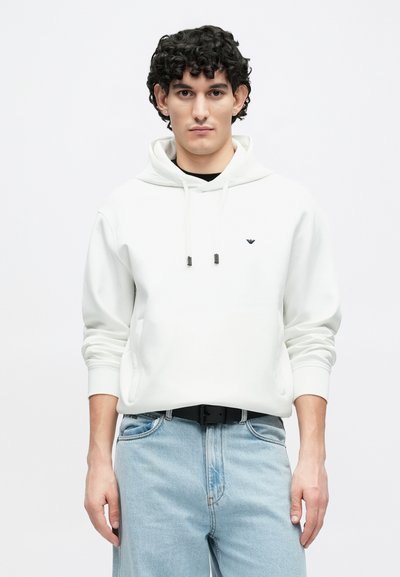 Witte hoodie met een voorvak en trekkoorden, voorzien van een klein zwart logo. Gecombineerd met lichtblauwe jeans en een zwarte riem.