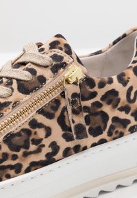 Sneaker en daim à motif léopard avec un détail de zip doré, des lacets beiges et une semelle plateforme blanche avec des bords texturés.