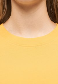 Κοντινό πλάνο του λαιμού και του πάνω μέρους του στήθους ενός ατόμου που φοράει φωτεινό κίτρινο μπλουζάκι με στρογγυλή λαιμόκοψη, με μακριά καστανά μαλλιά ορατά στα πλάγια.