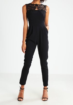 Mouwloze zwarte jumpsuit met kanten lijfje, aansluitende taille, taps toelopende broek en zwarte enkellaarzen met bandje, gedragen door een staande persoon.