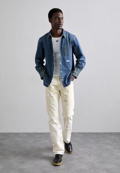 Camisa de denim con un solo bolsillo, combinada con pantalones blancos desgastados. Zapatillas negras con suela de goma completan el atuendo.