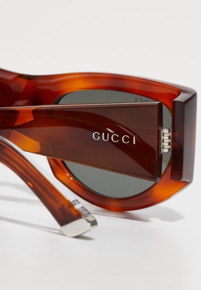 Gucci napszemüveg, fényes borostyán teknős pánttal, sötét lencsékkel és gravírozott logóval a szárán. Szögletes dizájn erős csuklós rendszerrel.