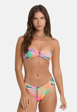 Młoda kobieta z długimi brązowymi włosami, ubrana w kolorowy, wzorzysty bikini bandeau i turkusowy naszyjnik, pozująca na jednolitym jasnym tle.