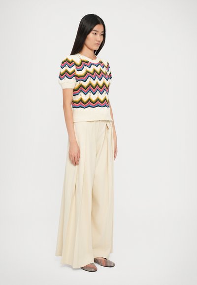 Haut en tricot crème à manches courtes avec un motif zigzag coloré, associé à un pantalon large de couleur beige clair et des chaussures plates rayées.