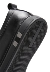 Scarpa nera slip-on con suola in gomma testurizzata. Il materiale superiore è in pelle liscia, con un anello posteriore e accenti metallici decorativi.