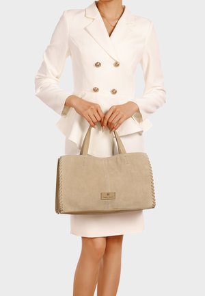 Donna con blazer bianco doppiopetto e gonna che tiene una grande borsa beige in suede con lati cuciti e due manici.
