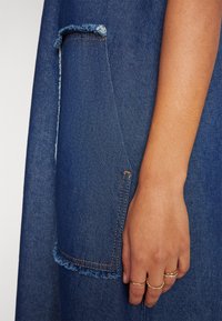 Denim-mekko, jossa on räjähtänyt tasku, sininen väri oransseilla ompeleilla, ja lähikuva kädestä, jossa on useita sormuksia.
