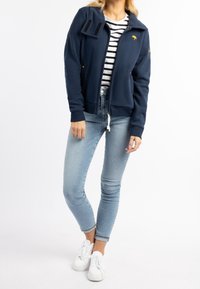 Felpa con zip di colore blu navy e colletto alto, dotata di una tasca a toppa, dettagli dorati e una texture morbida, abbinata a jeans skinny blu chiaro e sneaker bianche.