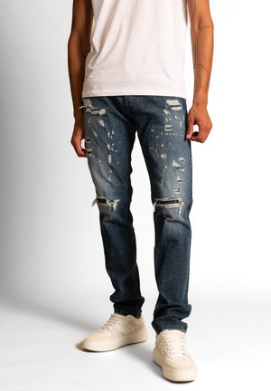 Mann trägt dunkelblaue zerrissene Jeans, weiße Sneakers und ein schlichtes weißes T-Shirt, steht vor einem einfarbigen weißen Hintergrund.