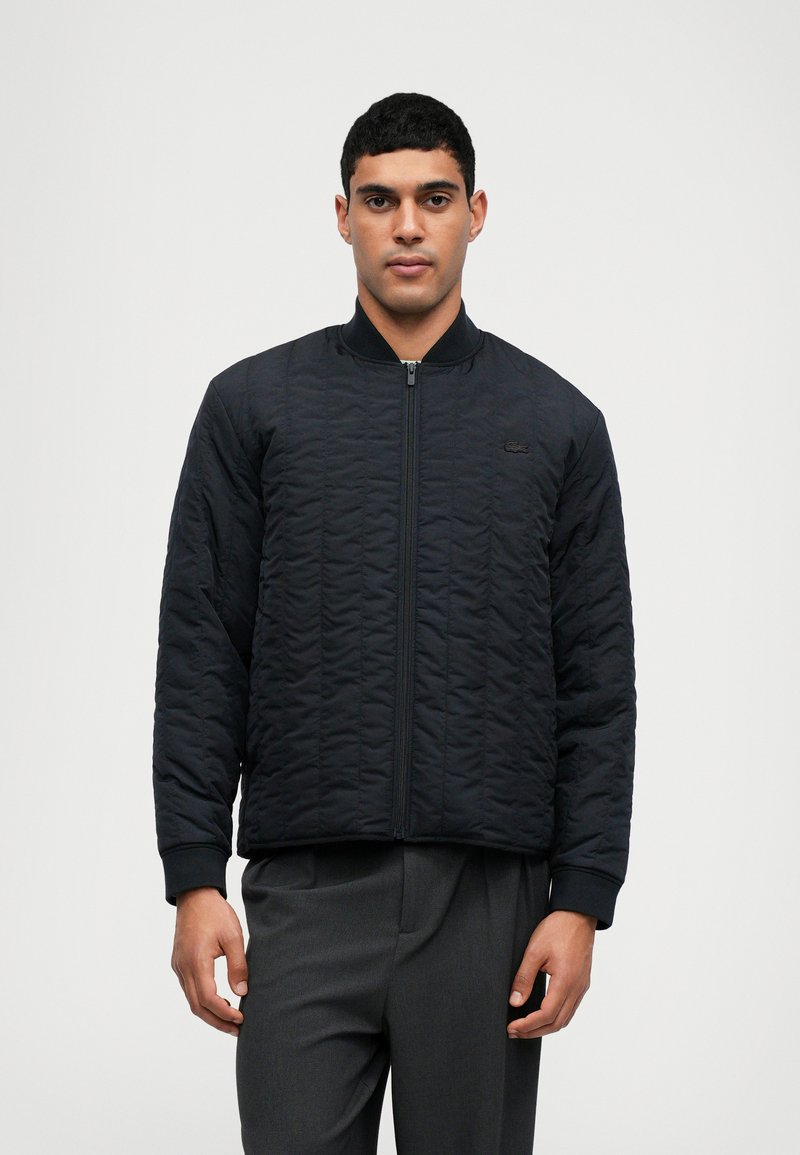 Blouson bomber matelassé bleu foncé avec fermeture zippée, poignets côtelés et un logo discret sur la poitrine. Tissu lisse et texturé.