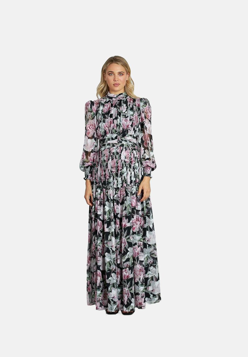 Vrouw in een lange, doorzichtige zwarte jurk met een roze en witte bloemenprint, hoge hals, opgeblazen mouwen en geplooide rok, staande met haar gezicht naar voren.